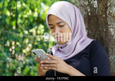 Close-up shot of Muslim dame en hijab en utilisant smartphone avec l'écouteur. Avec des jeunes Malaisiens femme ou student looking at smartphone. Banque D'Images