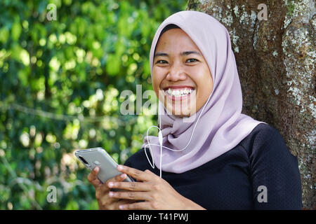 Close-up shot of Muslim dame en hijab en utilisant smartphone avec l'écouteur. Avec des jeunes Malaisiens femme ou student looking at smartphone. Banque D'Images