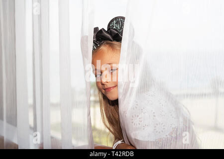 La petite fille est assise sur la fenêtre et à la recherche de derrière les rideaux.La petite fille est assise sur la fenêtre. Banque D'Images