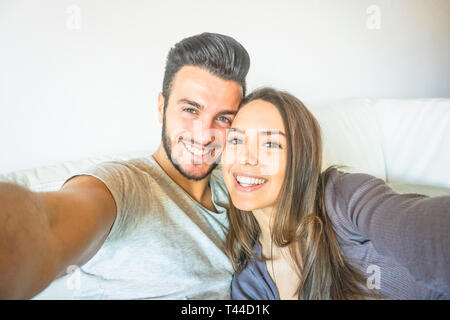 Happy young couple en tenant un téléphone intelligent selfies avec caméra dans le salon embracing on sofa at home - Amis self portrait Banque D'Images