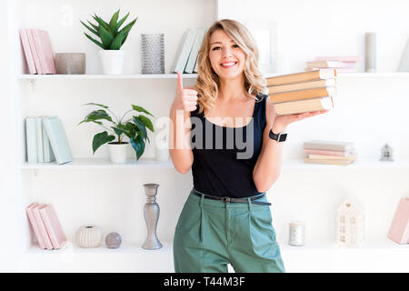 Belle fille blonde tenant une pile de livres et showing Thumbs up. Le concept de l'éducation, cours, examens, devoirs, liste de livres à lire Banque D'Images