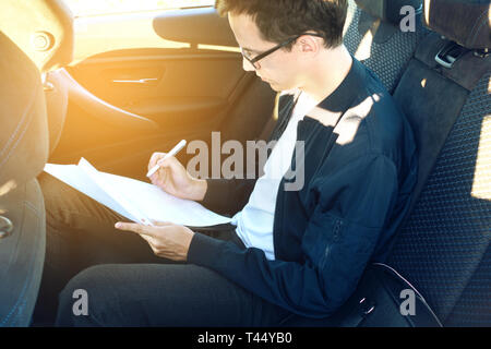Jeune homme d'affaires dans les verres signe les documents assis dans le siège arrière de la voiture. tonique Banque D'Images