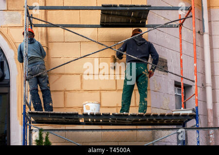 Deux travailleurs dans un casque et des vêtements de construction sur un escalier élevé sont la peinture avec des pinceaux et rouleaux, la réparation de la façade de la maison Banque D'Images