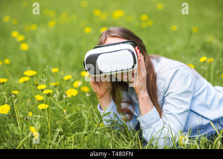 Femme à l'aide du casque de réalité virtuelle à l'extérieur pose en champ de fleurs de printemps Banque D'Images
