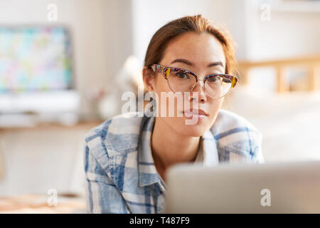 Portrait of Contemporary Asian woman wearing glasses using laptop lying on lit dans la lumière du soleil et à la recherche à l'appareil photo, copy space Banque D'Images
