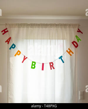 Image de la guirlande de lettres Happy Birthday pendants devant une fenêtre avec rideau blanc Banque D'Images