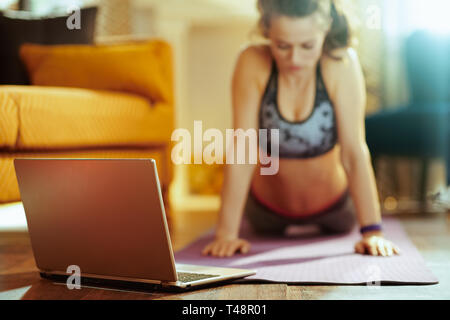Gros plan sur ordinateur portable beige et la femme en arrière-plan qui s'étend sur le tapis de fitness remise en forme tout en regardant des vidéos sur internet dans la maison moderne. Banque D'Images