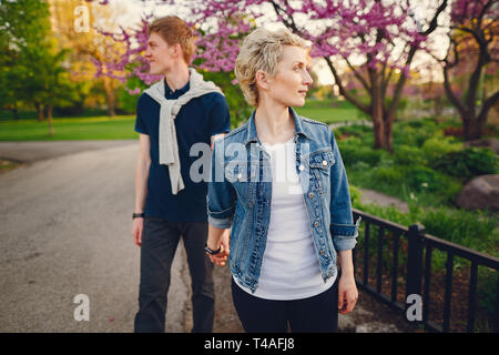 Femme élégante et belle avec les cheveux courts, vêtu d'une veste bleu jeans promenades avec son bel homme dans un parc ensoleillé et près de standind flower Banque D'Images