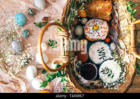 Panier de Pâques élégant moderne avec des œufs, du pain, du jambon gâteau de Pâques, des betteraves, des saucisses, de beurre et de branches vertes sur tissu rustique avec des fleurs de printemps dans la région de sunn Banque D'Images