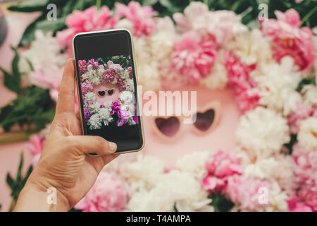 Main tenant le téléphone et la prise de photo de pink heartshaped sunglassess dans l'élégant quartier de pivoines rose et blanc cadre sur du papier rose télévision lay. Blogging Instagram Banque D'Images