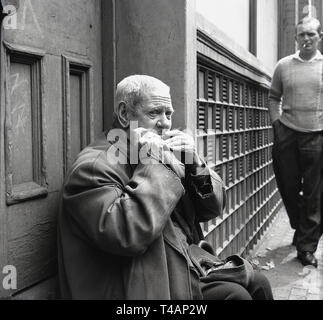 Années 1960, historique, un musicien ambulant, un homme âgé assis dans une rue porte avec son bonnet ouvert sur ses genoux, la lecture d'un ou de l'harmonica à bouche par Stepney marché, East London, Londres, Banque D'Images