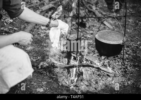 La reconstitution médiévale habillé en allemand de la Seconde Guerre mondiale soldat de la Wehrmacht à la cuisson des aliments sur un feu dans un vieux pot en marche. Photo en noir et blanc. Soldat Banque D'Images