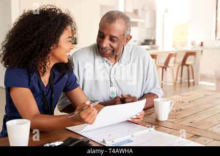 Happy female healthcare worker sitting at table smiling with a senior man au cours d'une visite à domicile Banque D'Images