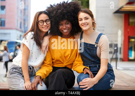 Trois jeunes adultes branchés copines sur un banc dans une rue de ville souriant pour appareil photo, Close up Banque D'Images