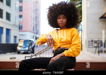 Jeune femme noire avec les cheveux afro et jaune haut assis sur une chaise dans la rue à la recherche d'appareil photo Banque D'Images