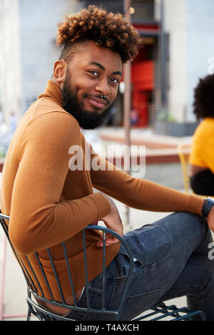 Hipster noir millénaire homme assis à l'extérieur d'un café dans la rue, se retourne en souriant à la caméra, Close up Banque D'Images