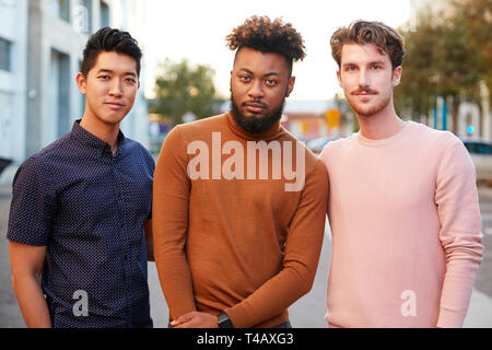 Les jeunes adultes de sexe masculin de la hanche trois amis debout dans une rue de la ville à la recherche d'appareil photo, vue de face, Close up Banque D'Images