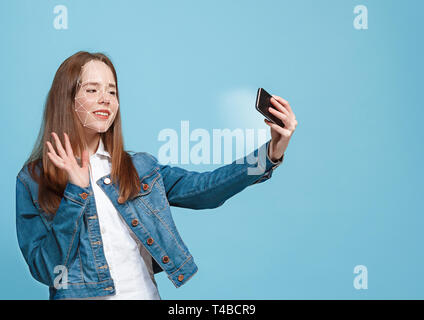 Essayer quelque chose de nouveau. Une fille avec un smartphone en jeans fonction debout sur fond bleu. La technologie de la reconnaissance faciale sur grille polygonale. Concept de cyber sécurité, affaires, travail, éducation. Banque D'Images