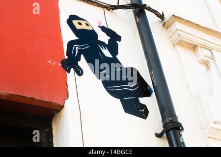 Graffiti par James AME - AME72 (le 'Guy Lego) - sur un mur à Aberystwyth montrant un noir adapté 'ninja' personnage apparemment tomber d'une gouttière tenant un peut de pulvérisation de peinture après peinture un coeur rose sur le mur. 10 mars 2019 Banque D'Images