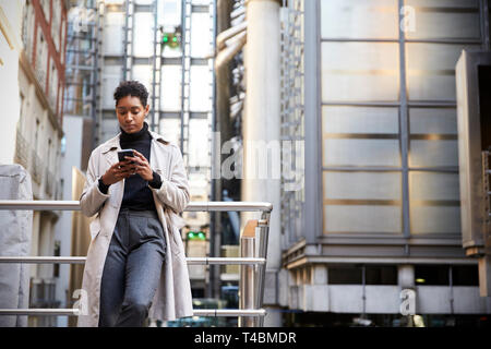 Jeune femme noire à la mode dans la ville debout appuyée sur une main courante à l'aide de son smartphone, low angle Banque D'Images