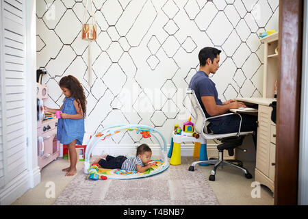 Papa est assis à un bureau de travail à la maison pendant que son petit fils et sa jeune fille jouer dans la pièce derrière lui Banque D'Images