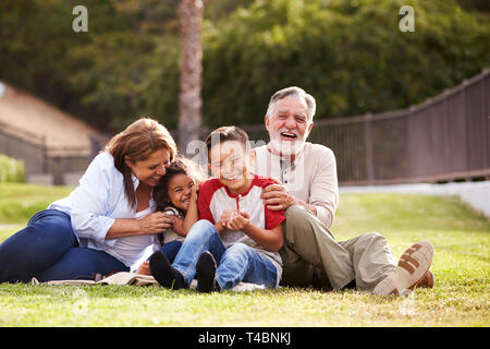 Les grands-parents hispaniques assis sur l'herbe dans le parc avec leurs petits-enfants rire, low angle Banque D'Images