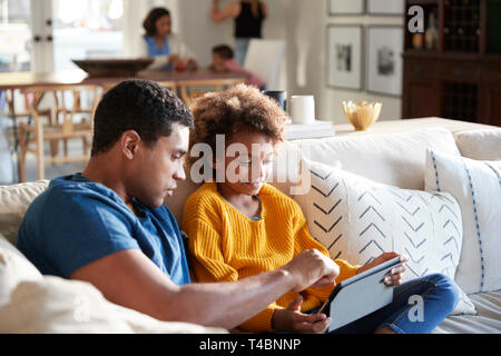 Pre-teen girl sitting on sofa dans le salon à l'aide de l'ordinateur tablette avec son père, mère et enfant assis à une table dans l'arrière-plan, selective focus Banque D'Images