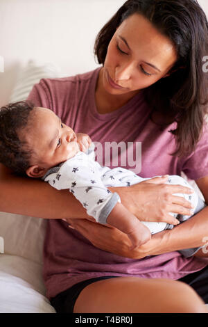 Mixed Race millénaire mère assise sur son lit tenant son bébé de trois mois fils dans ses bras, vertical Banque D'Images