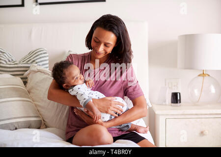 Mixed Race millénaire mère assise sur son lit tenant son bébé de trois mois fils dans ses bras Banque D'Images