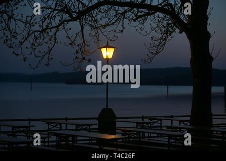 Manche, en Allemagne. Apr 15, 2019. Tables et bancs d'un jardin de la bière sont à l'heure bleue sur la rive de l'Ammersee sous une lanterne. Credit : Karl-Josef Opim/dpa/Alamy Live News Banque D'Images