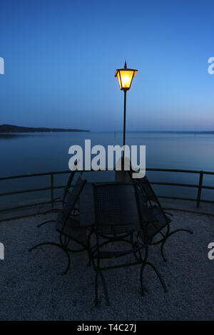 Manche, en Allemagne. Apr 15, 2019. Tables et chaises d'un café en plein air sont à l'heure bleue sur la rive de l'Ammersee sous une lanterne. Credit : Karl-Josef Opim/dpa/Alamy Live News Banque D'Images