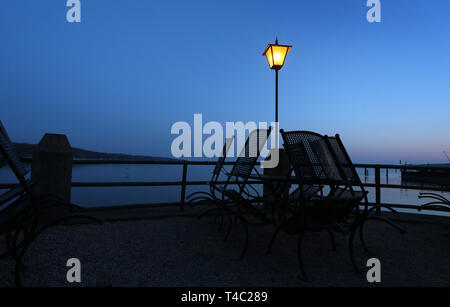 Manche, en Allemagne. Apr 15, 2019. Tables et chaises d'un café en plein air sont à l'heure bleue sur la rive de l'Ammersee sous une lanterne. Credit : Karl-Josef Opim/dpa/Alamy Live News Banque D'Images