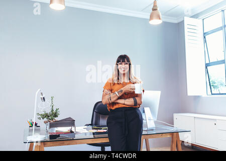 Smiling woman entrepreneur debout près de son tableau de l'office de tenir une tasse de café. Cheerful businesswoman dans son bureau prendre une tasse de café. Banque D'Images