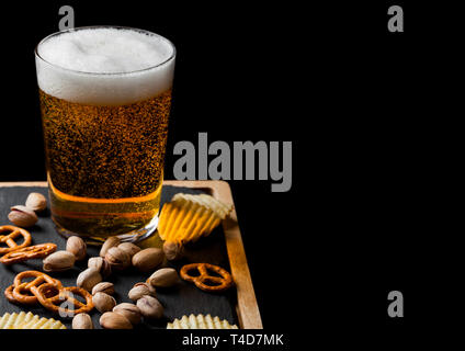 Verre de bière blonde avec un en-cas sur le conseil de pierre sur fond noir. Pistaches et bretzel avec chips de pommes de terre Banque D'Images