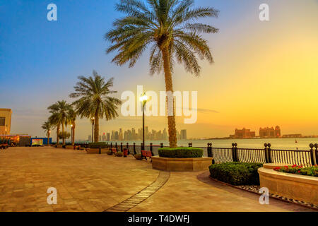 Des bancs et des palmiers le long passage à Porto marina Saoudite au Pearl-Qatar, Doha, avec des gratte-ciel de West Bay skyline illuminé de heure bleue Banque D'Images