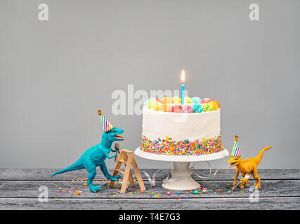 Dinosaures jouets faim port d'un chapeau et tenant fourches à côté d'un gâteau d'anniversaire sur fond gris Banque D'Images