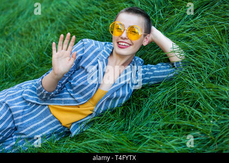 Portrait de la belle jeune femme aux cheveux courts en costume rayé bleu décontracté, chemise jaune verres couché sur l'herbe verte, à la recherche à camea avec t Banque D'Images