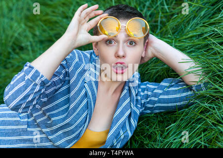 Portrait de jeune femme mignon surpris avec les cheveux courts en costume rayé bleu couché sur l'herbe verte, hoding verres jaunes à Banque D'Images