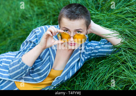 Portrait de jeune femme avec des cheveux courts en costume rayé bleu décontracté, chemise jaune et les lunettes couché sur l'herbe verte et à la recherche à Banque D'Images