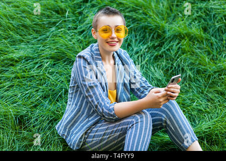 Portrait de jeune femme cheveux courts en costume rayé bleu, verres jaunes sitting on grass tenant son smart phone mobile et à la recherche en c Banque D'Images