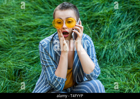 Portrait of young woman in casual cheveux courts costume rayé bleu, chemise jaune et verres assis sur l'herbe verte parlant au téléphone et surp Banque D'Images