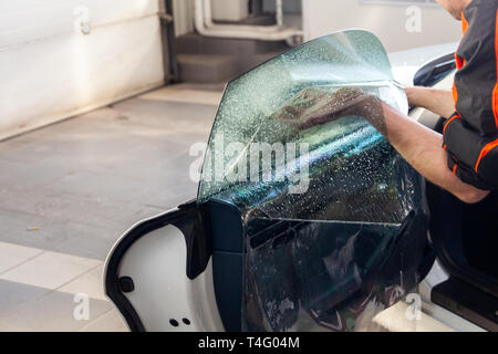 L'assistant d'installation d'équipement supplémentaire bois une teinte film sur le côté de la vitre avant de la voiture et il s'aplatit à la main pour monter la vitre avec un g Banque D'Images