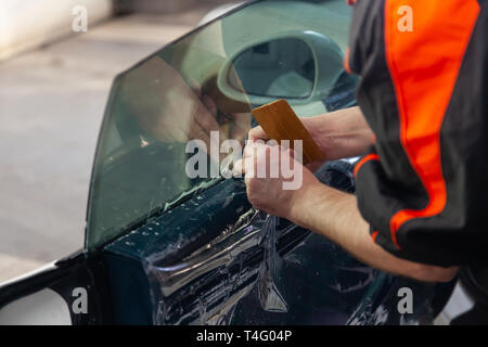 L'assistant d'installation d'équipement supplémentaire bois une teinte film sur le côté de la vitre avant de la voiture et il s'aplatit à la main pour monter la vitre avec un g Banque D'Images