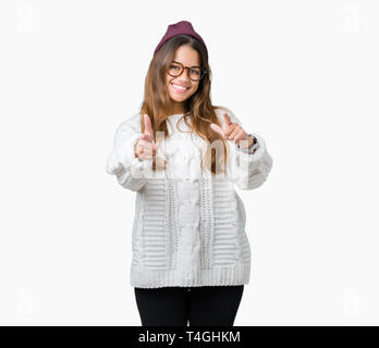 Belle jeune femme brune portant des lunettes et hipster hiver chapeau sur fond isolé pointer du doigt à la caméra avec plaisir et drôle de visage. G Banque D'Images