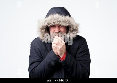 Homme mature avec barbe dans les vêtements d'hiver avec un capuchon sur la tête est la congélation, essayant de se réchauffer. Studio shot Banque D'Images