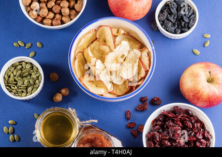 Tasse en céramique avec apple des croustilles maison entouré de blanc tasses avec des collations saines, les pommes fraîches et de petits pots de confiture et miel sur dos bleu vif Banque D'Images