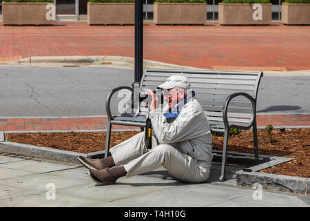 ASHEVILLE, NC, USA-4/11/19 : Un homme assis sur le sol devant un banc pour prendre des photos avec appareil photo Nikon, et souriant. Banque D'Images