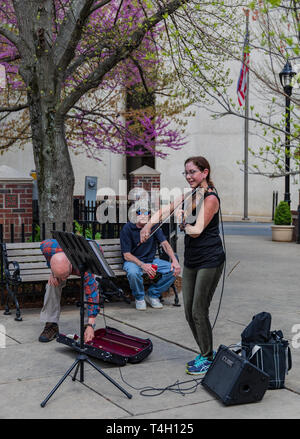 ASHEVILLE, NC, USA-4/11/19 : A woman smiling et arts de la rue au centre-ville, alors qu'un homme met l'argent dans son étui à violon, tandis qu'un autre homme s'assied et écoute. Banque D'Images