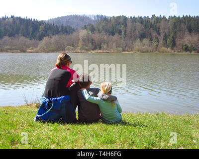 Mère avec son fils et sa fille assise sur une prairie d'herbe verte à la recherche au lac et forêt. Famille heureuse dans la nature Banque D'Images
