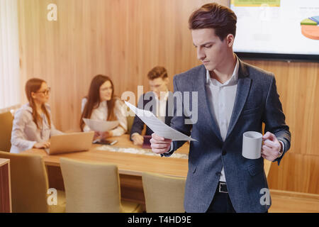 Un jeune homme élégant dans un veston et une chemise avec une tasse de café dans sa main se trouve et lit des documents sur le contexte de travail des collègues de l'off Banque D'Images